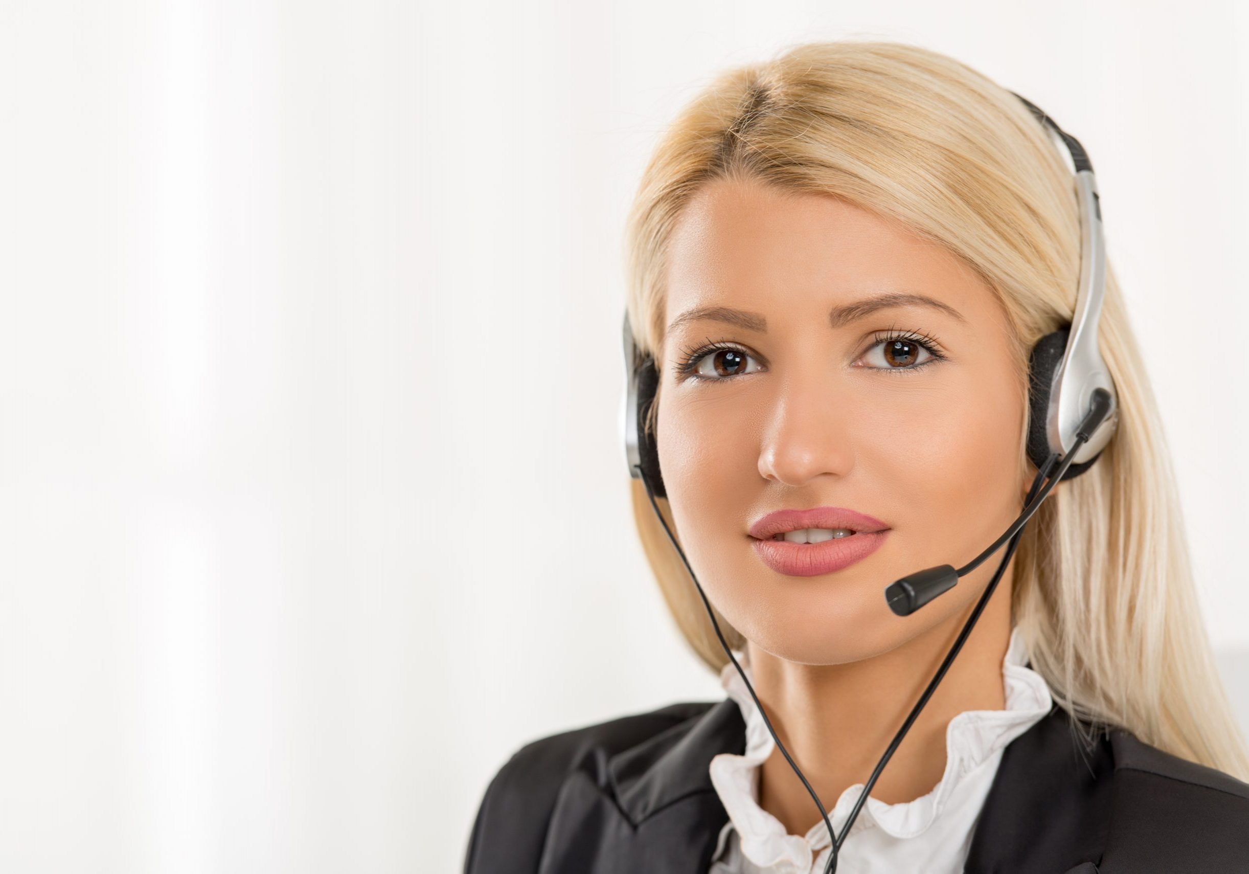 Close-up of a portrait young beautiful blonde girl elegantly dressed sitting at an office with a headset on her head. With a smile looking at the camera.